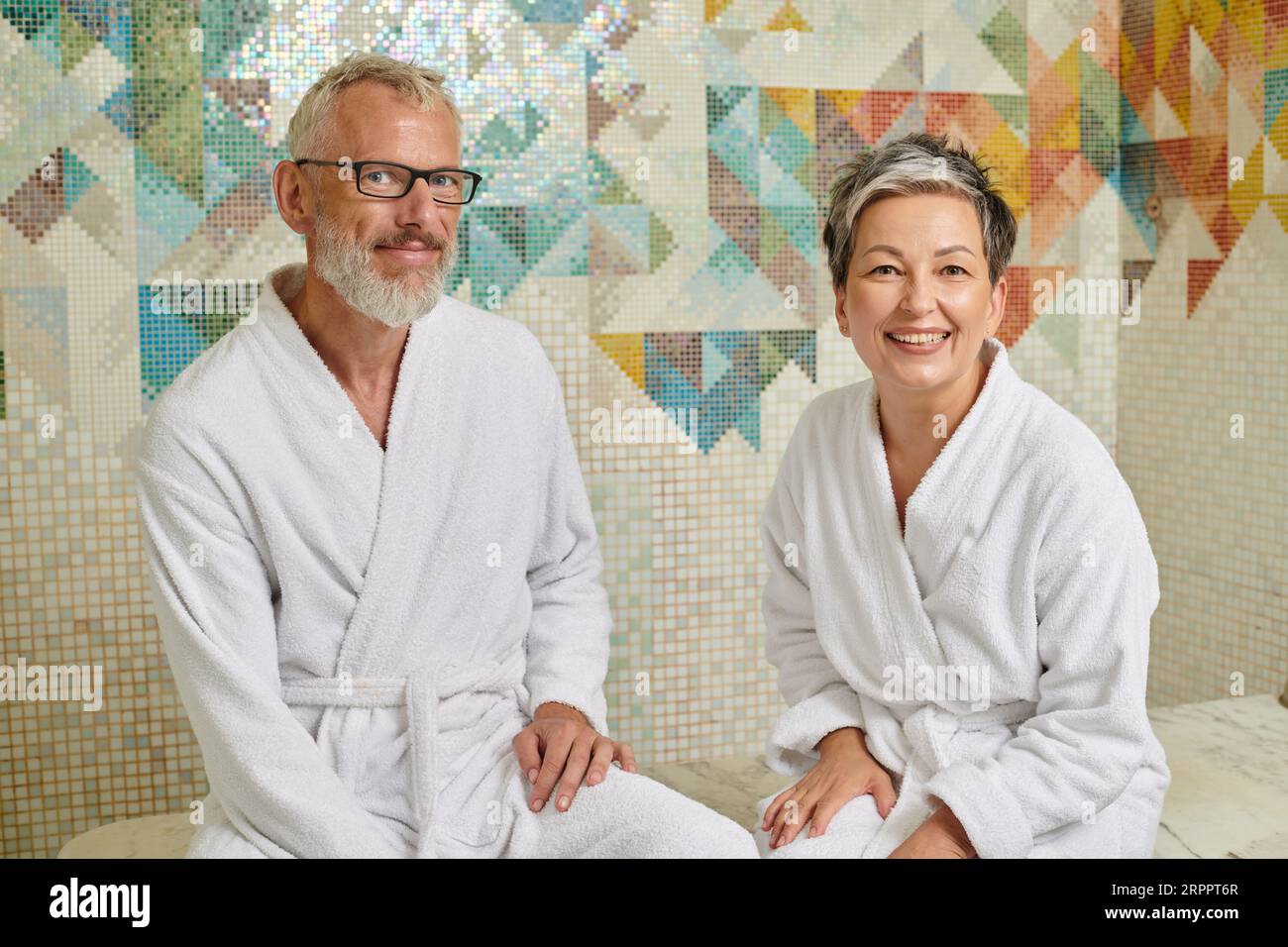 cheerful middle aged couple in white robes sitting in sauna, spa and wellness concept, hammam