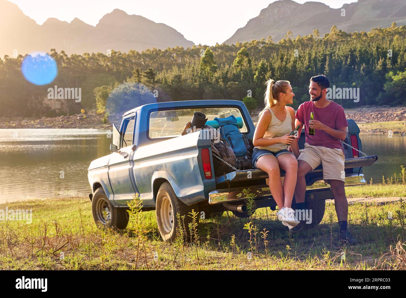 Couple With Backpacks In Pick Up Truck On Road Trip By Lake Drinking Beer Stock Photo