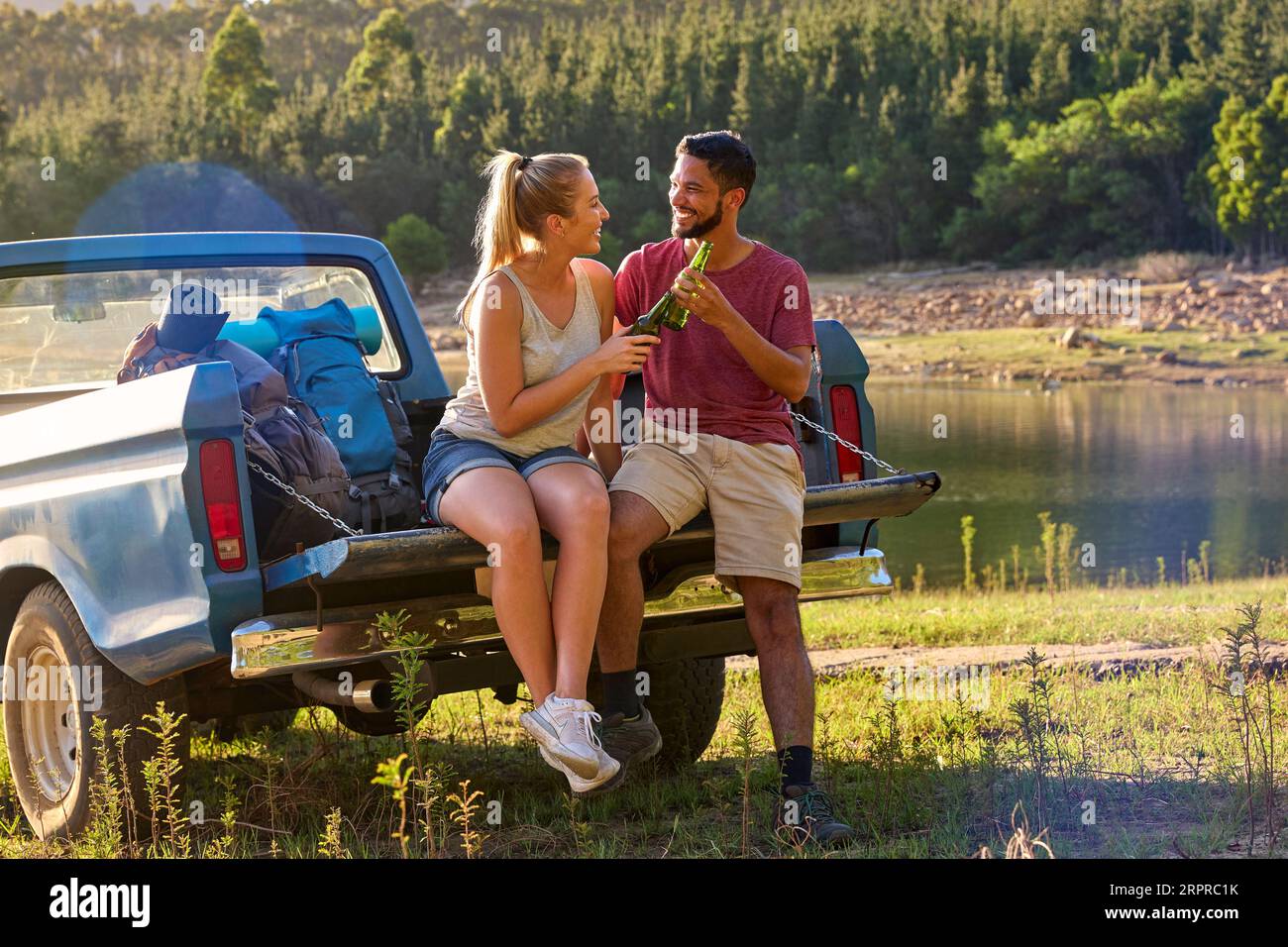 Couple With Backpacks In Pick Up Truck On Road Trip By Lake Drinking Beer Stock Photo