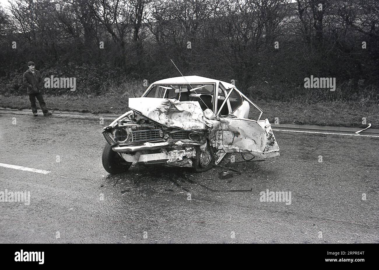 1983, historical, a man standing on the edge of a main road looking at his badly crashed car, having been involved in a head on collision on a wet, slippery moorland road, England, UK. Stock Photo
