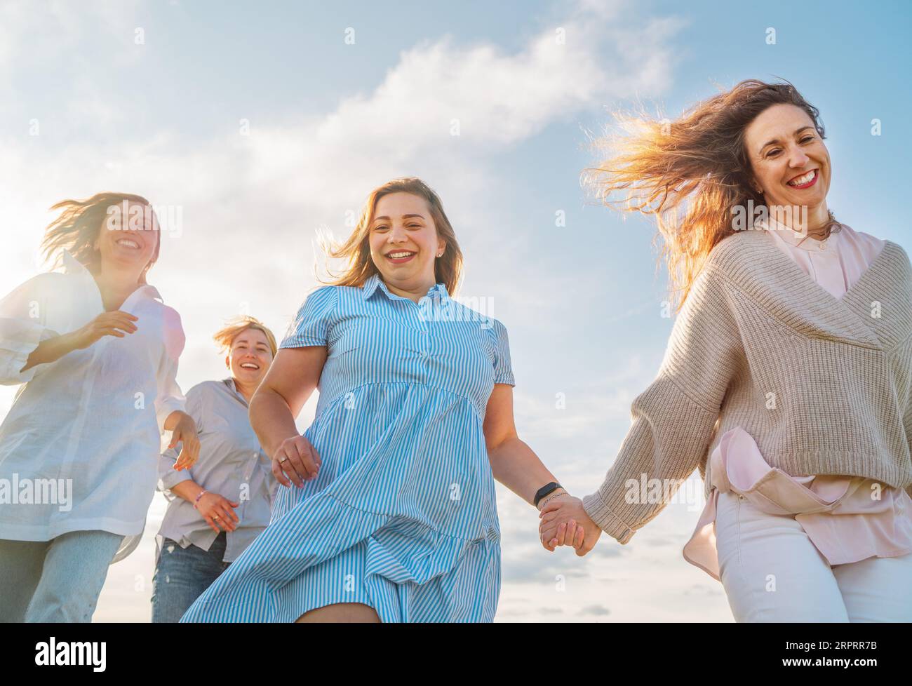 Portrait of four cheerful smiling women holding hand in hand running by the meadow. Low angle photo shot on the blue sky with clouds. Woman's friendsh Stock Photo
