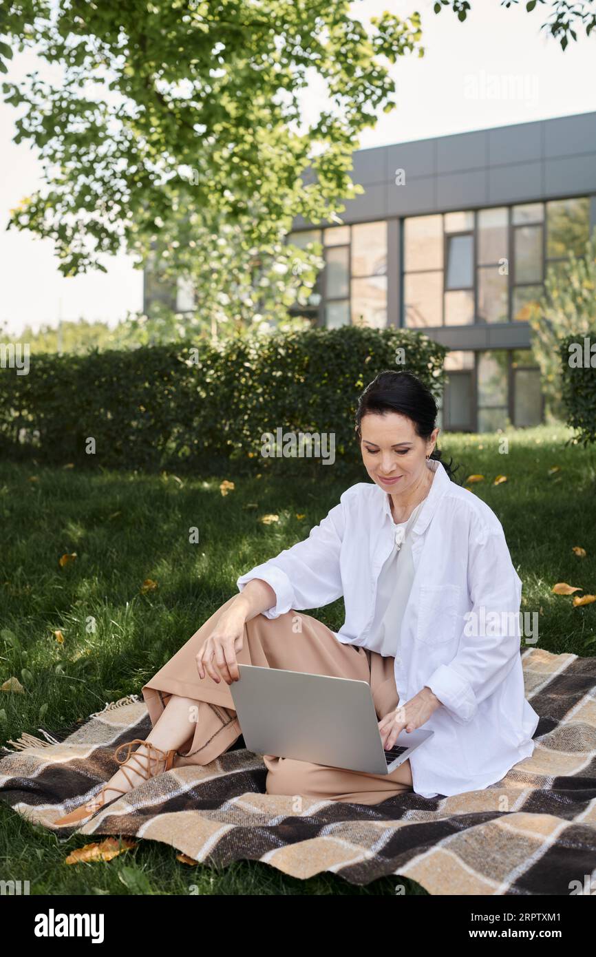 stylish and smiling middle aged woman working on laptop on blanket in park, work life balance Stock Photo