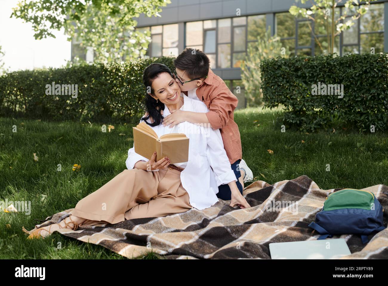 boy with down syndrome kissing happy mother reading book on blanket in park, emotional connection Stock Photo