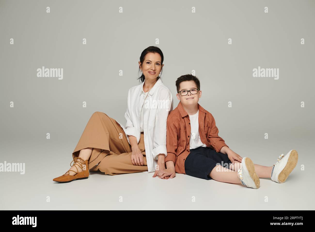 joyful boy with down syndrome and middle aged mother in stylish clothes smiling and sitting on grey Stock Photo