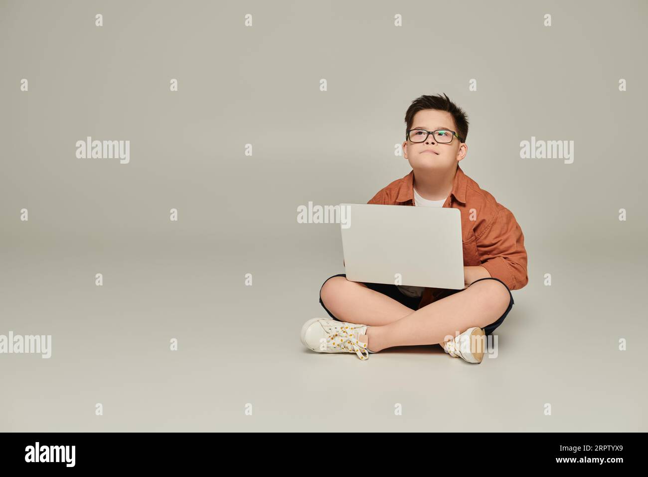 stylish and thoughtful boy with down syndrome sitting with laptop and crossed legs on grey Stock Photo