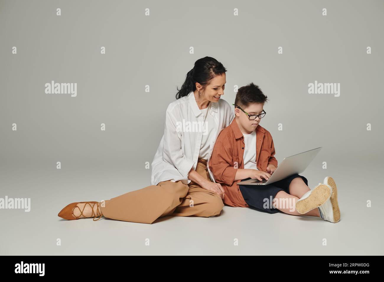 kid with down syndrome sitting near smiling mother and using laptop on grey, full length Stock Photo