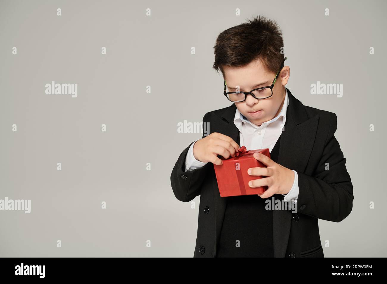 boy with down syndrome in school uniform and eyeglasses opening gift box with red ribbon on grey Stock Photo