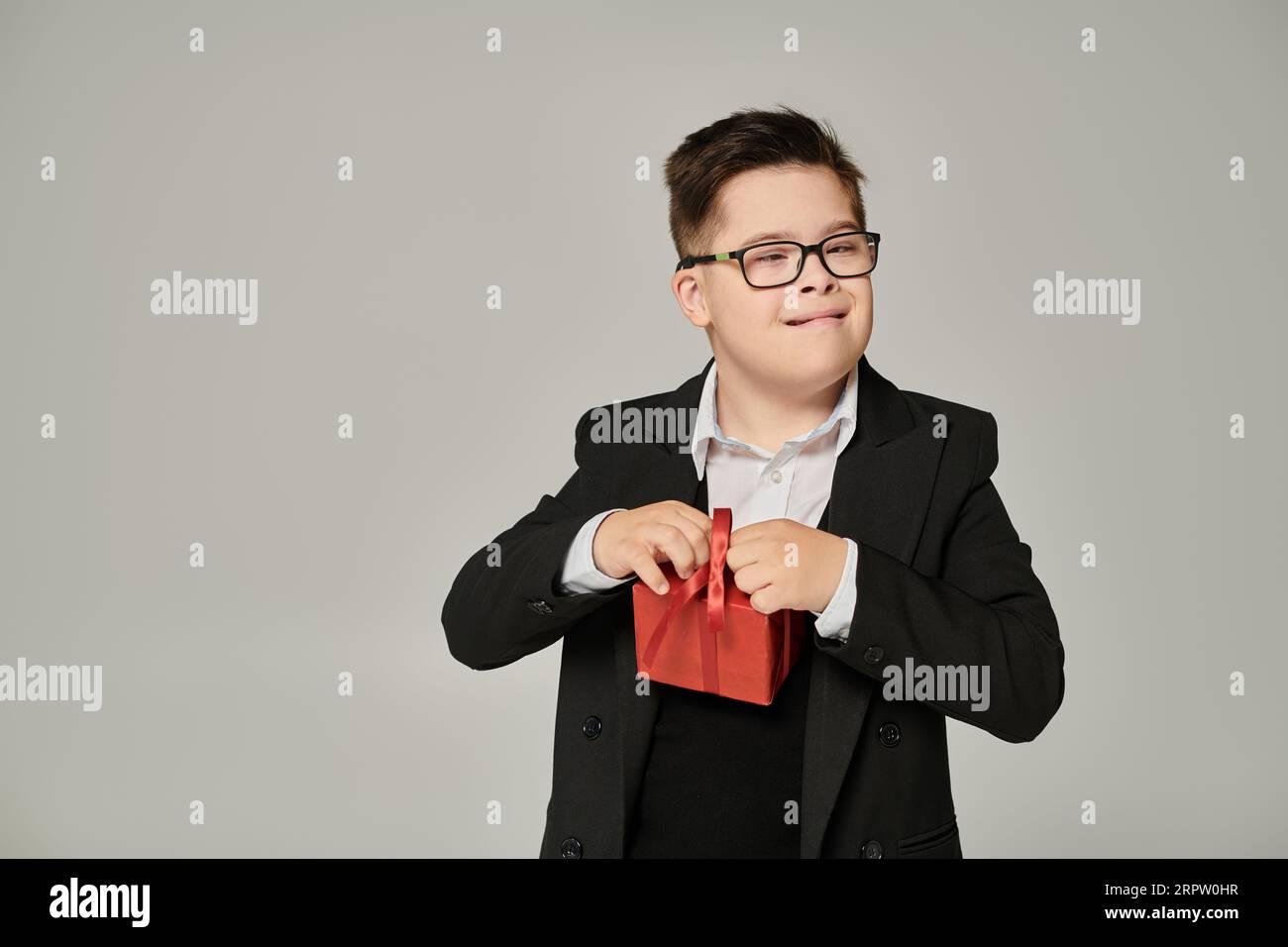 kid with down syndrome in eyeglasses and school uniform with gift box on grey, special student Stock Photo
