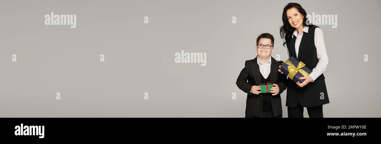happy special needs schoolboy with mother in formal wear holding gift boxes on grey, banner Stock Photo