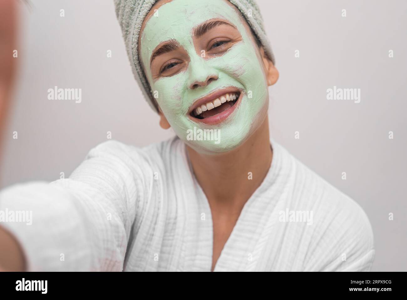 Joyful woman blogger with clay mask on face wearing bathrobe making selfie on white background Stock Photo