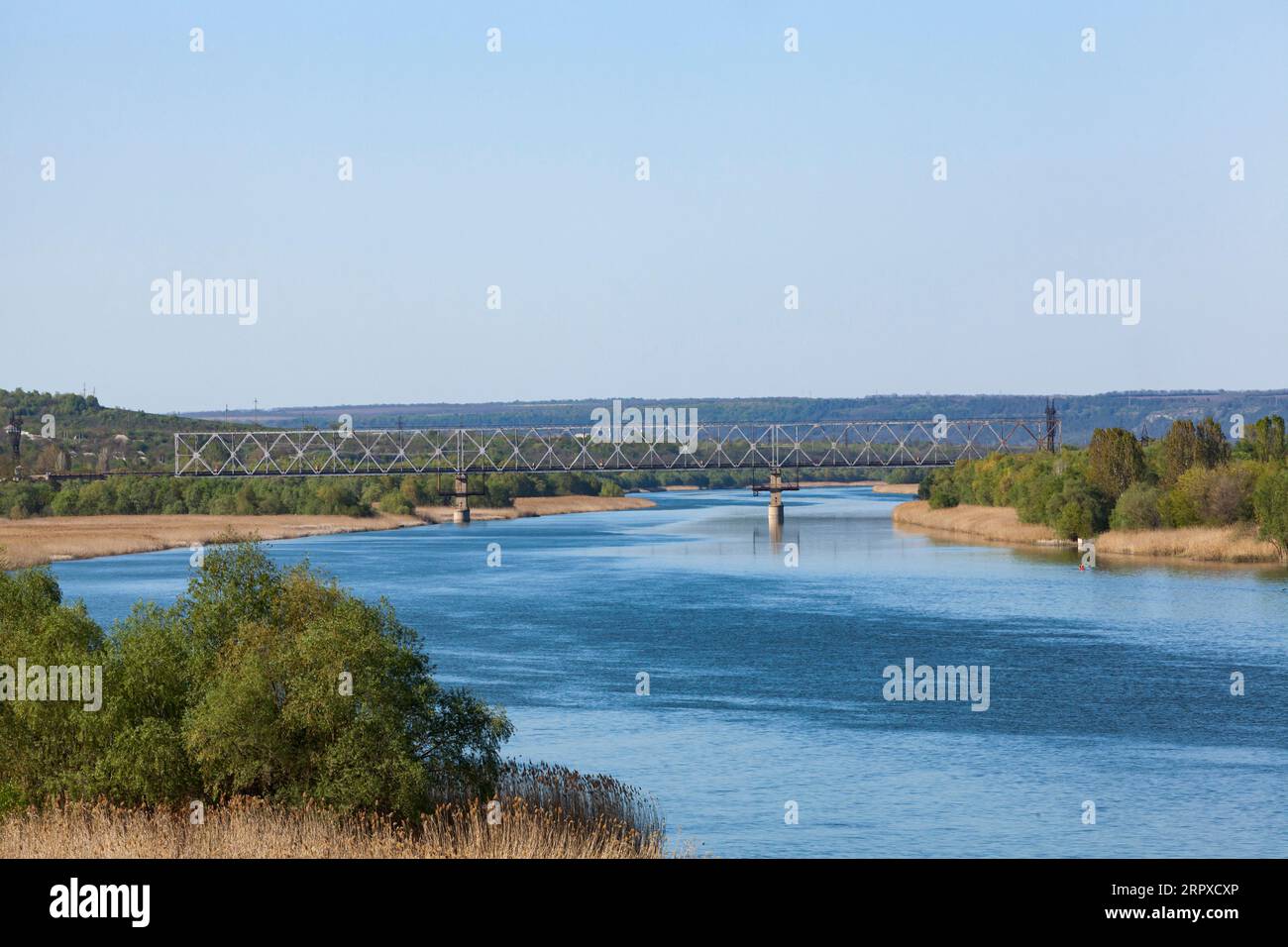 Dniester. View of the river from the railway bridge. The blue sky is reflected in the water. Green trees and dry reeds. Horizontal photo. Stock Photo