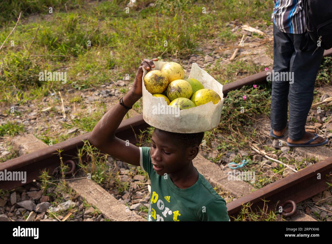 Boy Carrying Bucket of Monkey Orange Wild Fruits on his head Along the Railway Line Stock Photo
