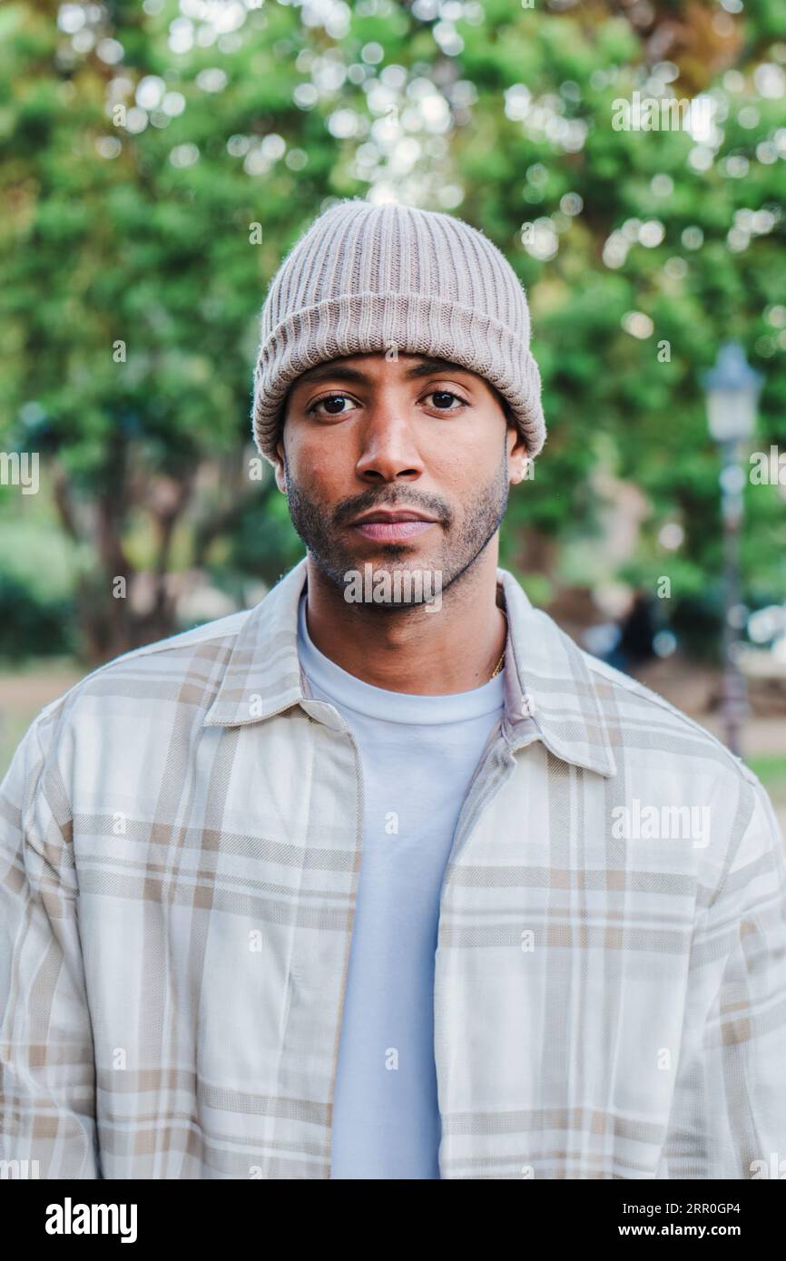 Vertical close up individual portrait of young attractive african american man with beanie hat looking serious at camera. Front view of a hispanic guy standing in a park outdoors with sad attitude. High quality photo Stock Photo