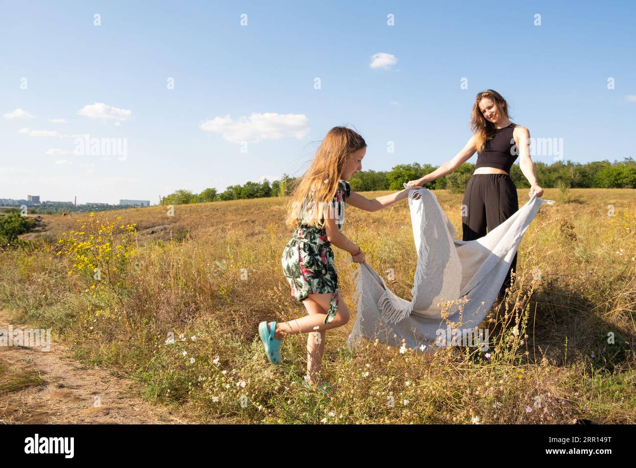 Mother and daughter are preparing for a picnic in the summer field, spreading a blanket on the grass Stock Photo