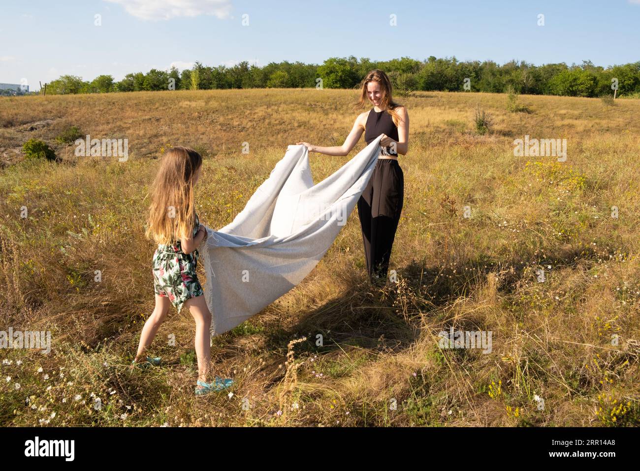 Mother and daughter are preparing for a picnic in the summer field, spreading a blanket on the grass Stock Photo