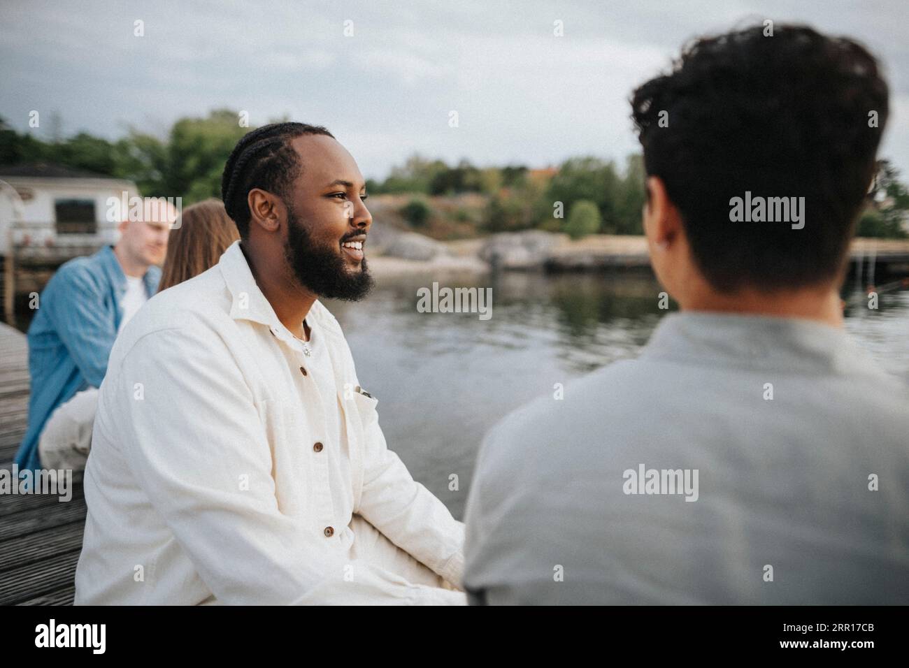 Smiling man looking away while sitting with friend on pier near lake Stock Photo