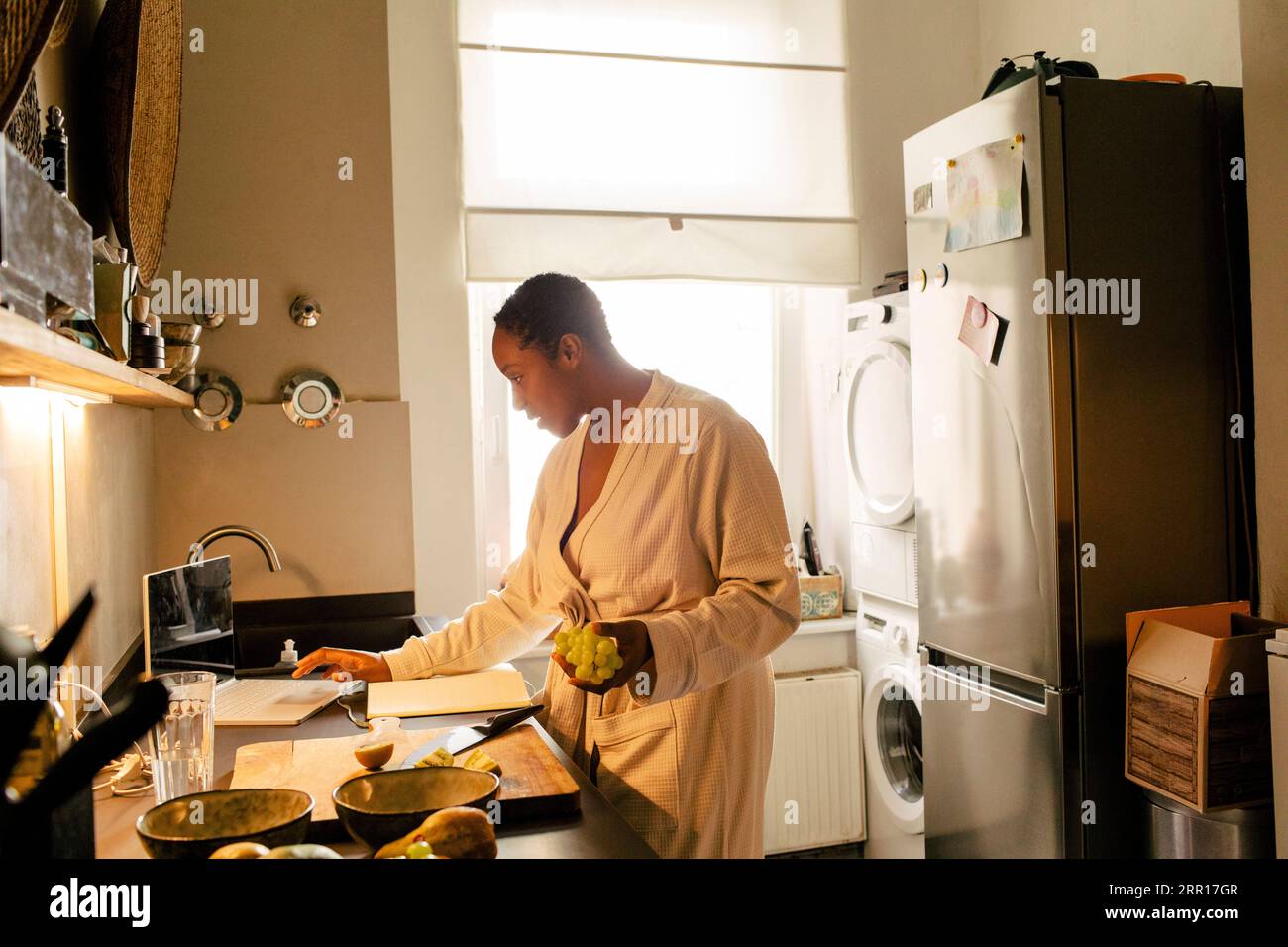 Young woman holding grapes using laptop in kitchen at home Stock Photo