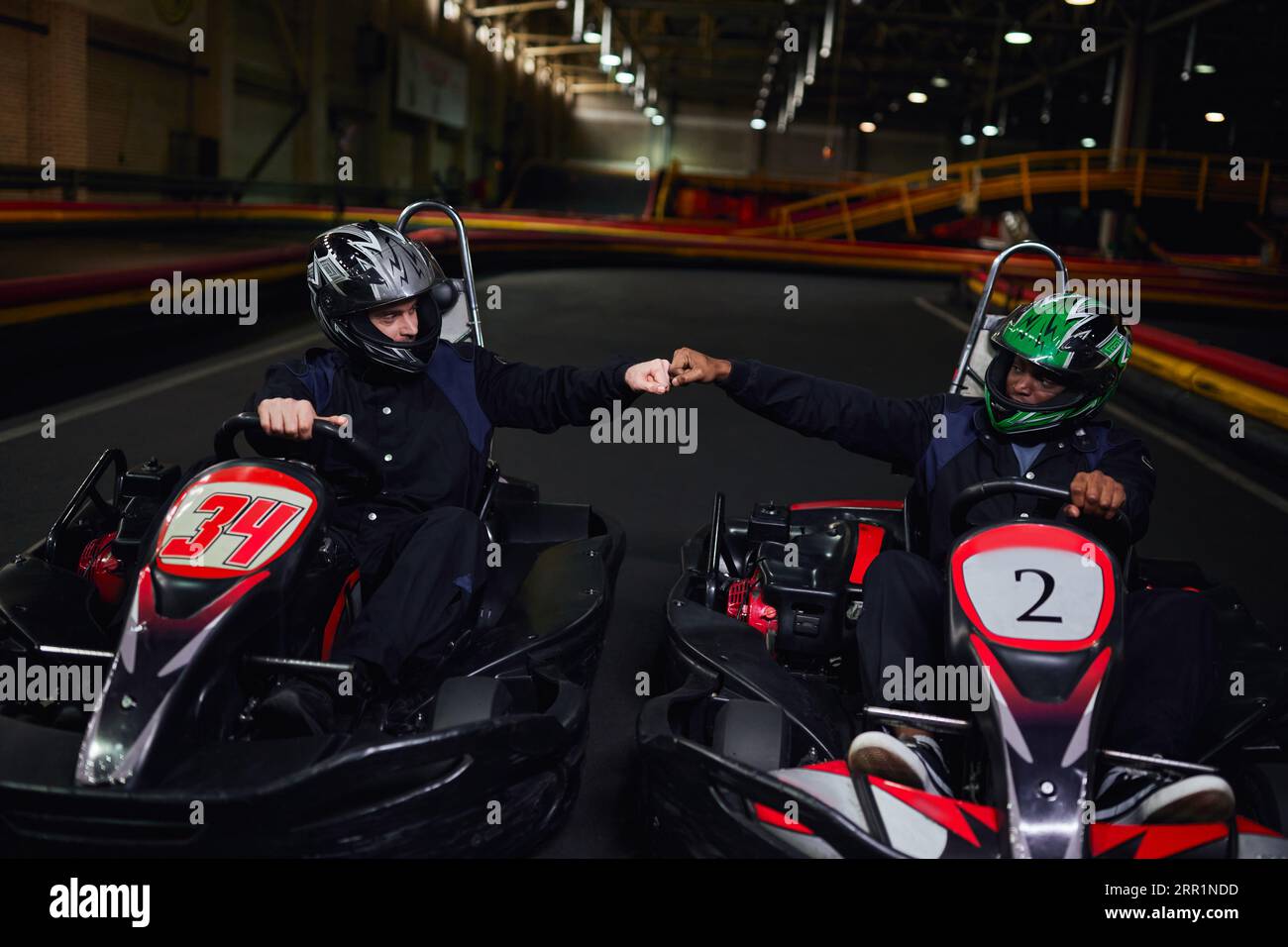 diverse go kart drivers in helmets fist bumping and sitting in sport cars for karting on circuit Stock Photo