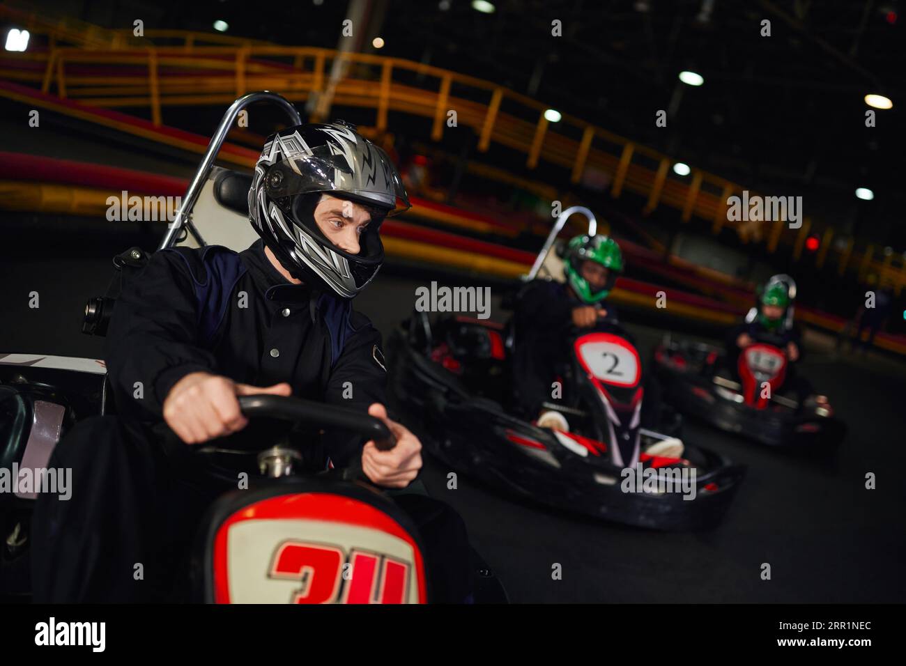concentrated man driving go kart near diverse drivers in helmets on indoor circuit, adrenaline Stock Photo