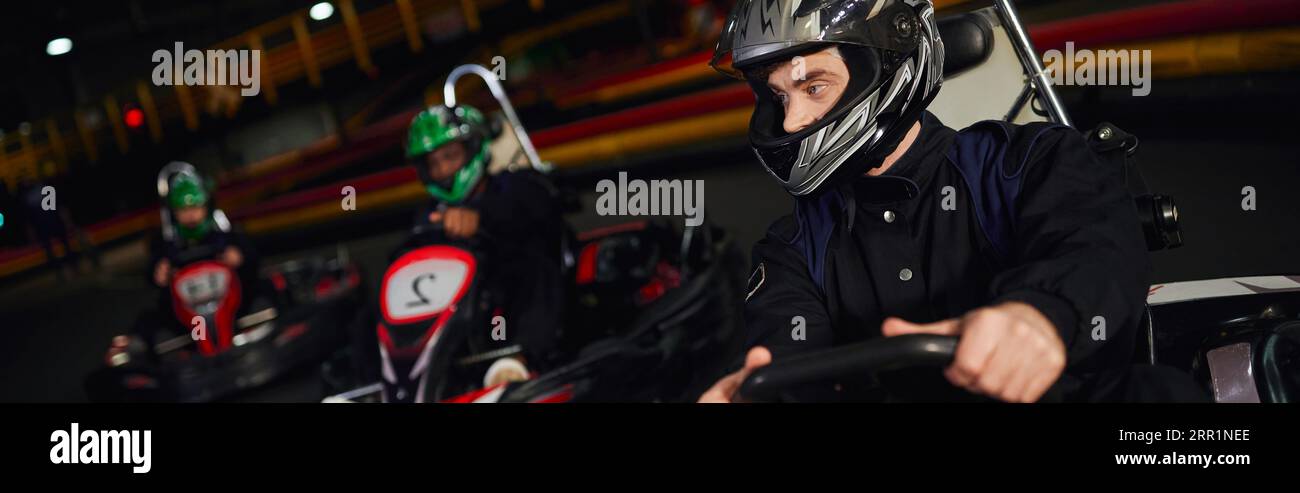 concentrated man driving go kart near diverse drivers in helmets on indoor circuit, banner Stock Photo