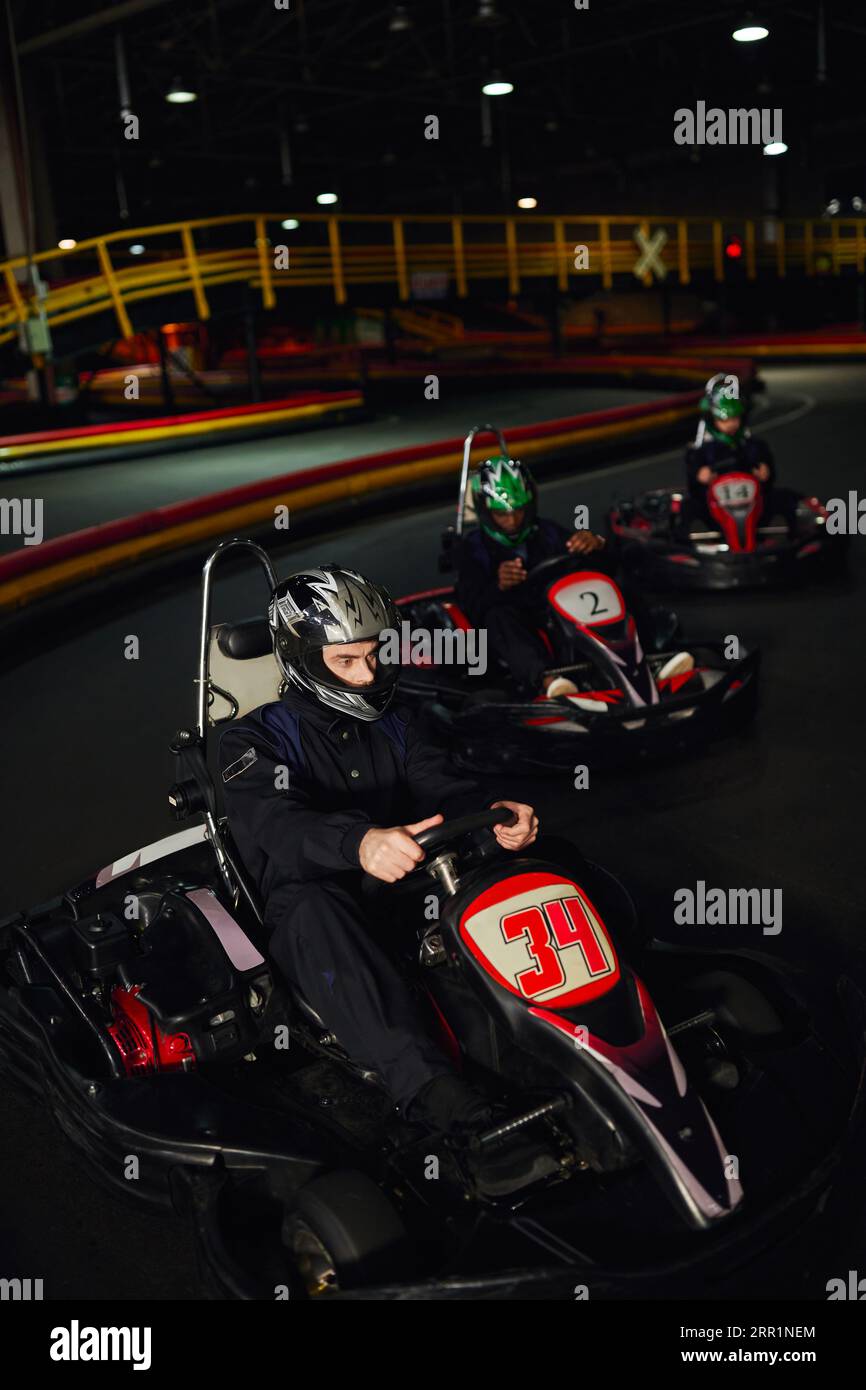 focused man driving go kart near diverse drivers in helmets on indoor circuit, adrenaline and sport Stock Photo
