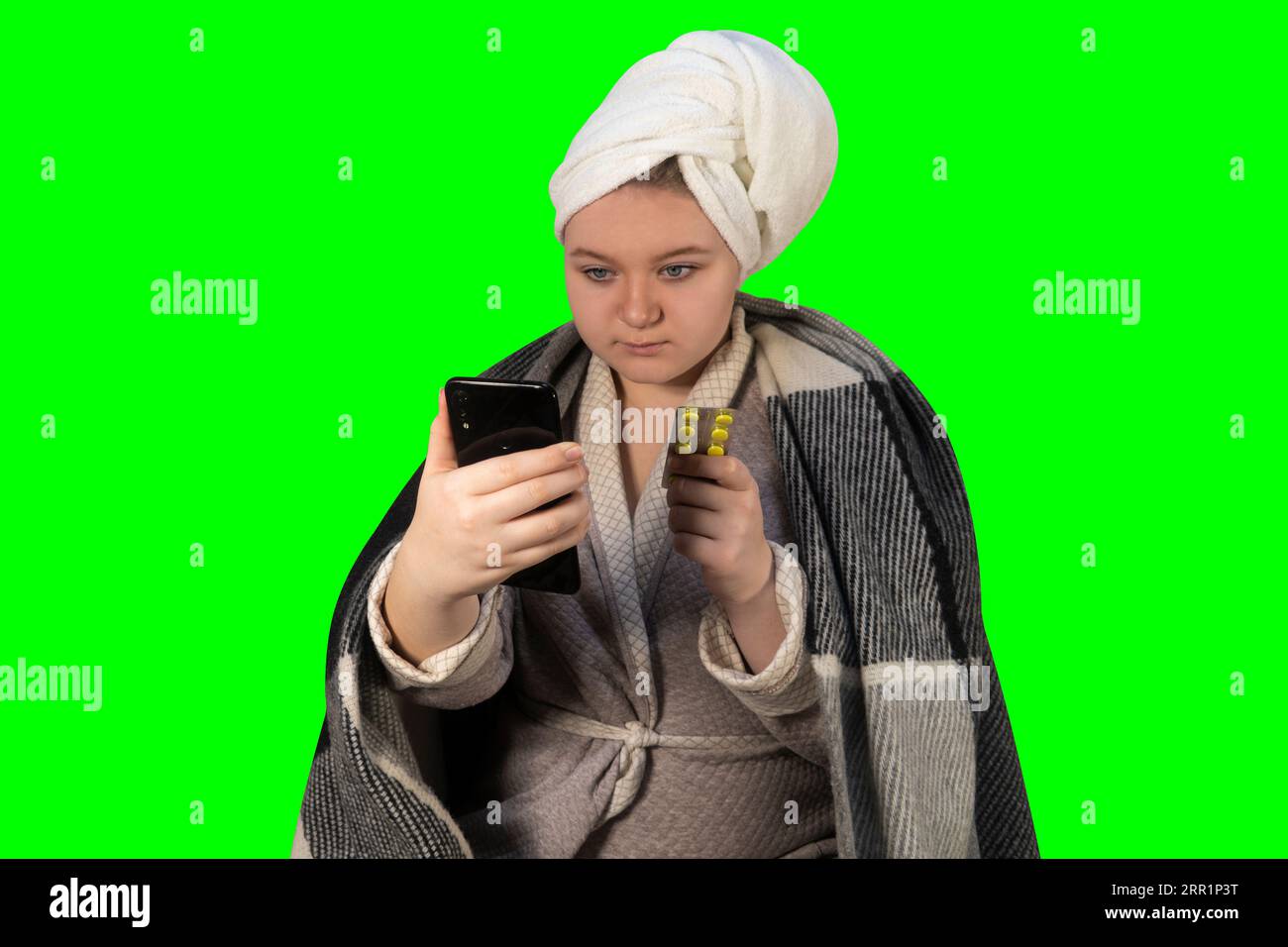 Girl with cold symptoms on a green background (chroma key) holds a blister of pills in one hand and a smartphone in the other. Online consultation Stock Photo