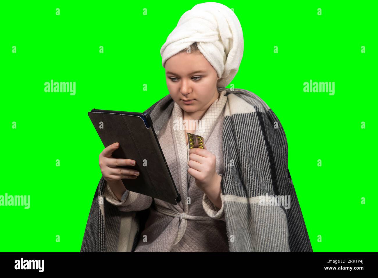 Girl with symptoms of cold holds a blister of pills in one hand and a tablet PC on a green background (chroma key) in other. Online consultation Stock Photo