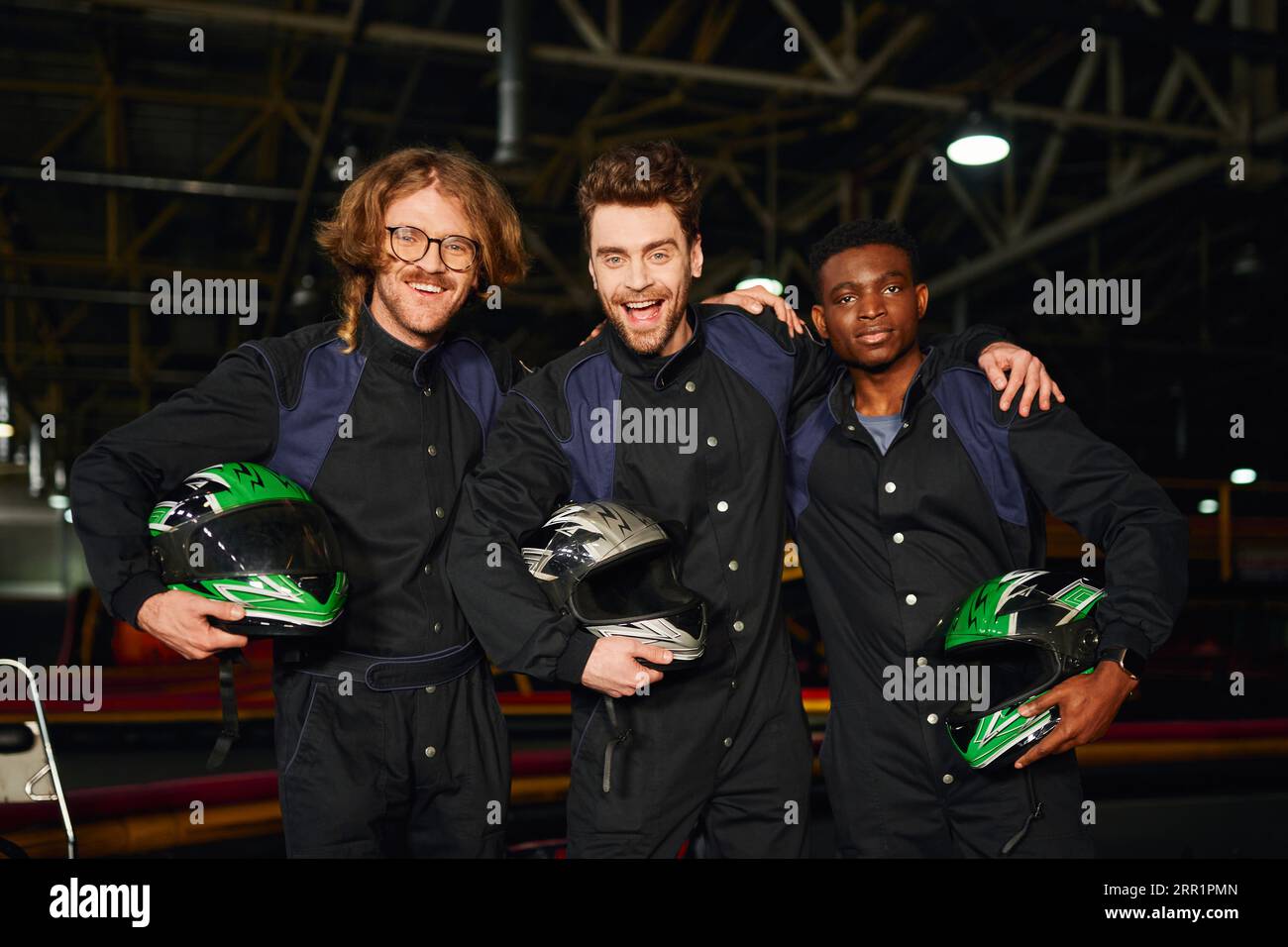 multicultural and excited go kart racers in protective suits hugging and holding helmets, winners Stock Photo