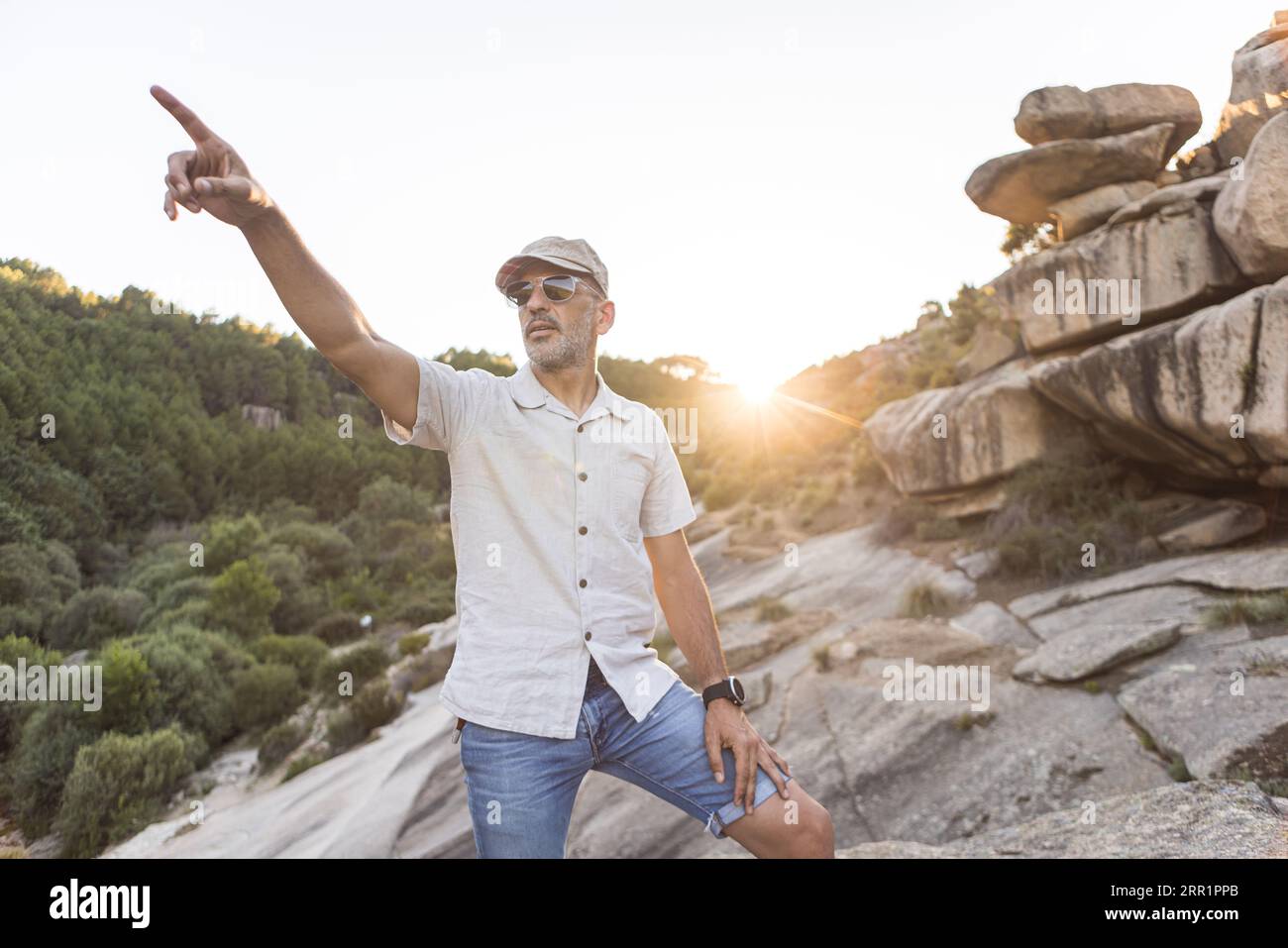 Serious bearded male in sunglasses and cap standing on rocky terrain near green forest in evening time and pointing aside Stock Photo