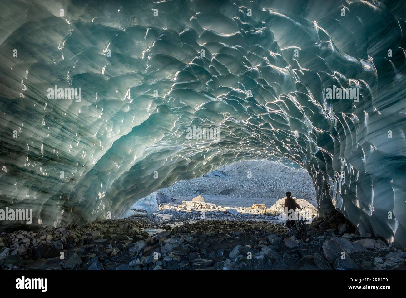 Back view of anonymous tourist standing near bike under hole while exploring ice cave in Switzerland on winter day Stock Photo