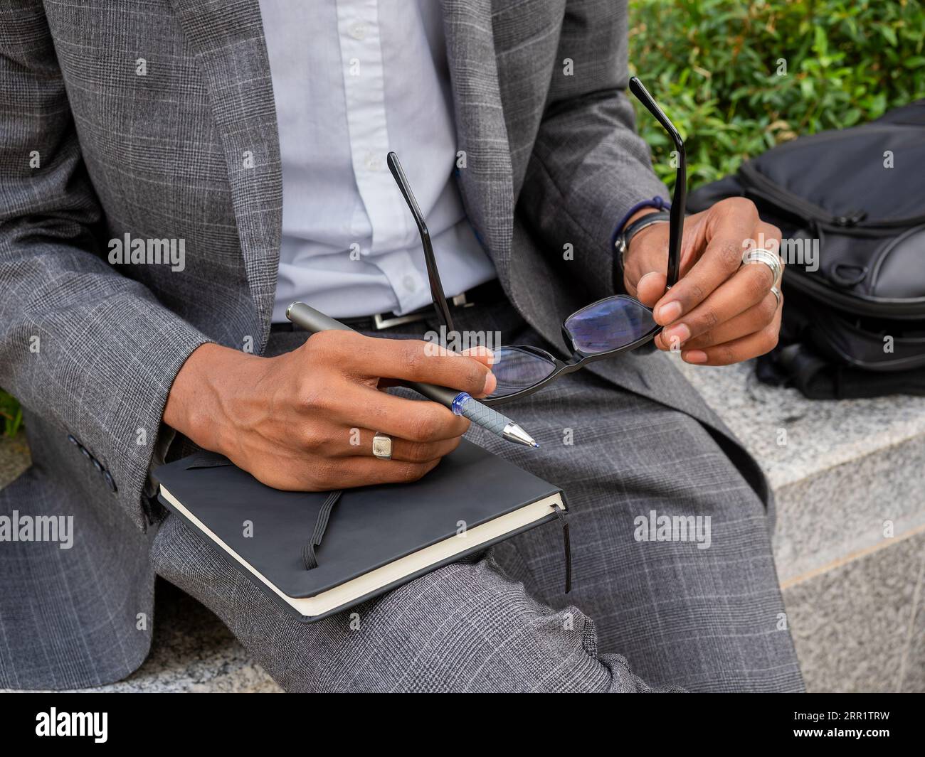 From above of anonymous black man in formal wear sitting on bench on street and holding eyeglasses and pen with notebook Stock Photo