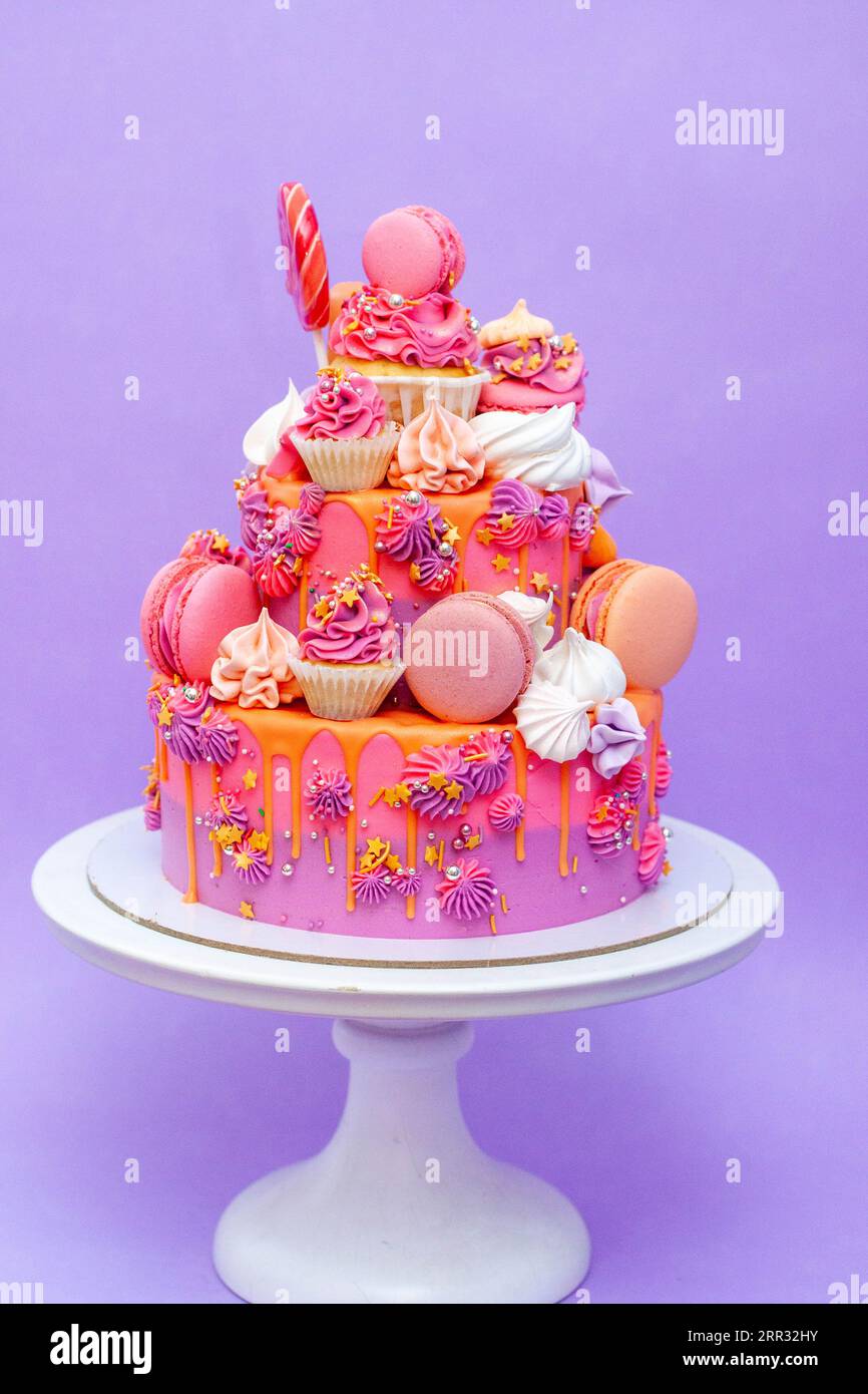 Pink, purple and orange gradient cake with cream cheese roses, macarons, cupcakes, meringues on purple plain background Stock Photo