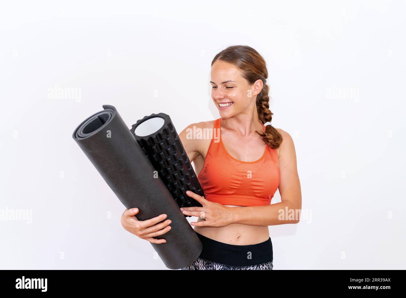studio shot female athlete holding exercise equipment foam roller rolled yoga mat and smiling, wearing sportswear, in front of white background. Stock Photo