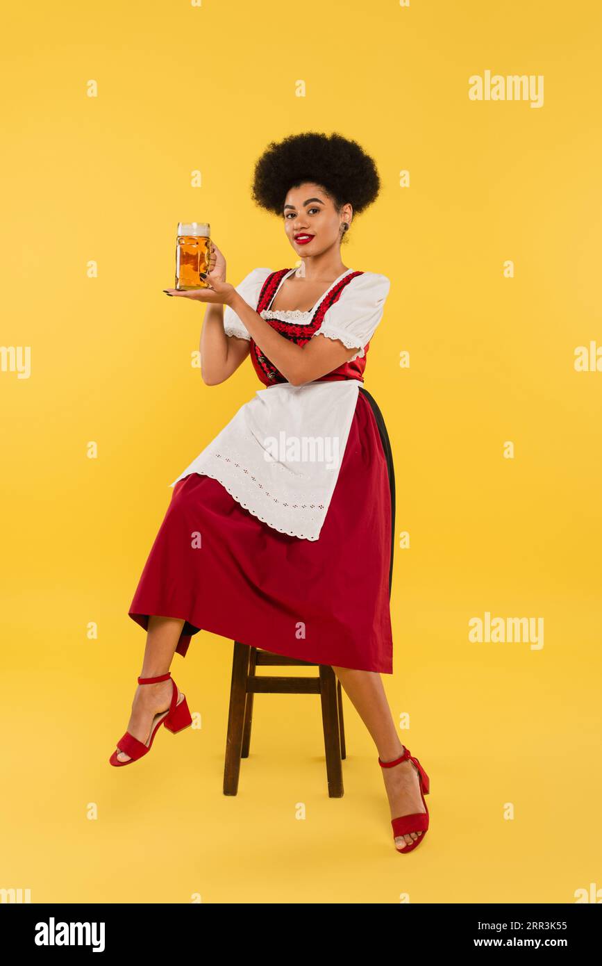 young african american waitress in authentic bavarian dress sitting on chair with beer mug on yellow Stock Photo