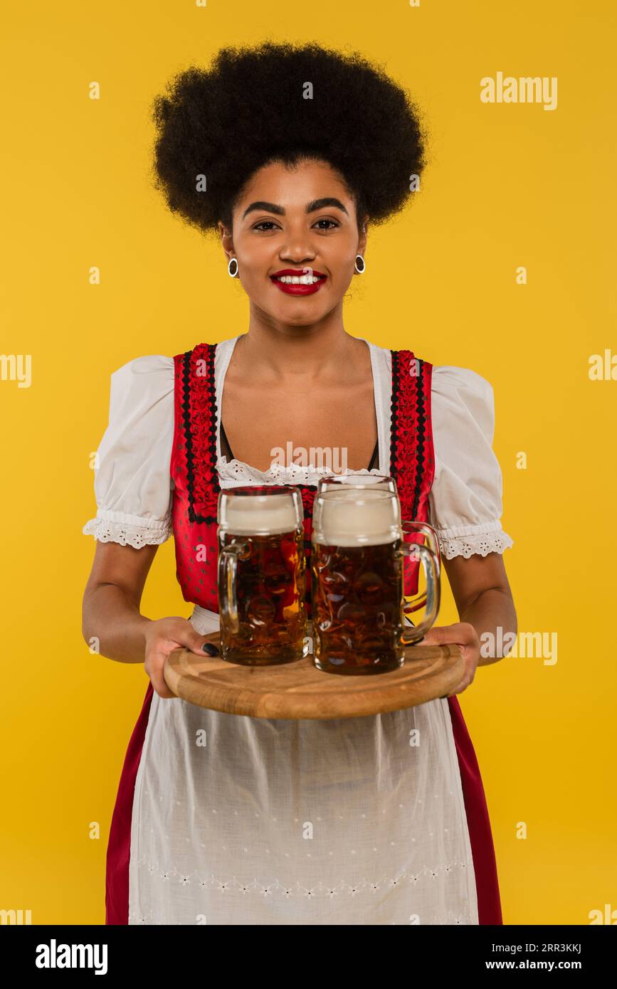 happy african american waitress in oktoberfest dress serving bavarian beer in mugs on tray on yellow Stock Photo