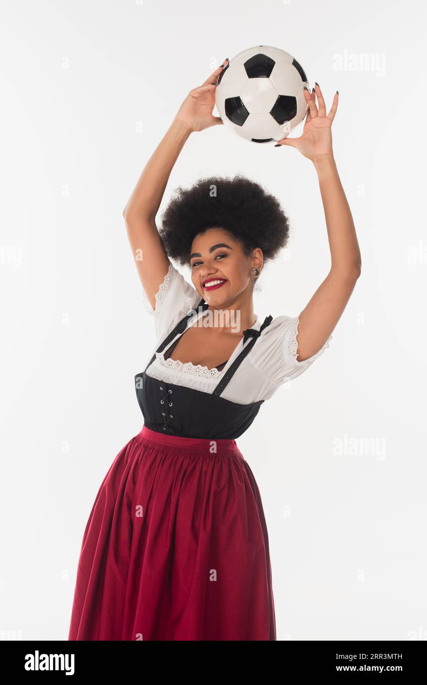 joyful african american bavarian waitress in dirndl holding soccer ball in raised hands on white Stock Photo