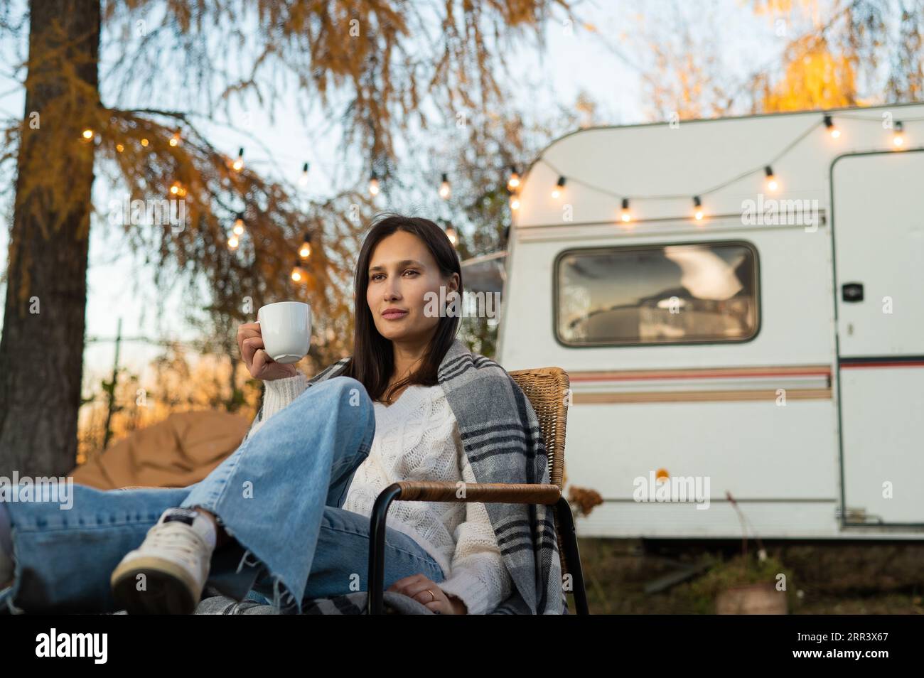 Caucasian woman in a knitted hat wrapped in a plaid and drinks a warming drink outdoors. Travel in a motor home in the fall. Stock Photo