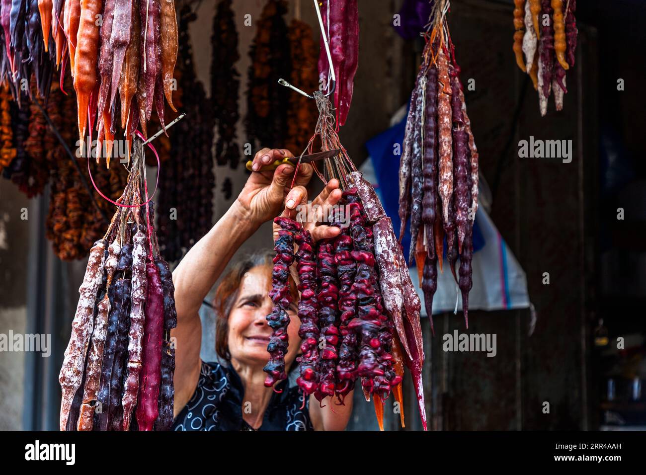 A vendor cuts a strand of tschurtschchela at her stall in the Dezerter Bazaar. Tschurtschchela (ჩურჩხელა) is a Georgian sweet: walnuts or hazelnuts are strung on threads and dipped in pelamushi, a coating made from reduced grape must and cornflour. The candle-shaped strands in red, purple and orange hang in dense clusters. Mikheil Tsinamdzghvrishvili Street, Chughureti, Didube, Tbilisi, 0102, Georgia Stock Photo