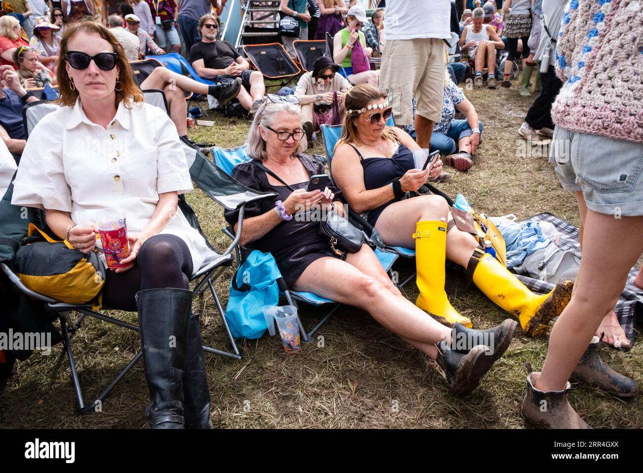 Two middle aged festival goers sitting in wellies in the mud scrolling on their mobile phones. Green Man Festival, Brecon, Wales, UK, 2023. Photo: Rob Stock Photo