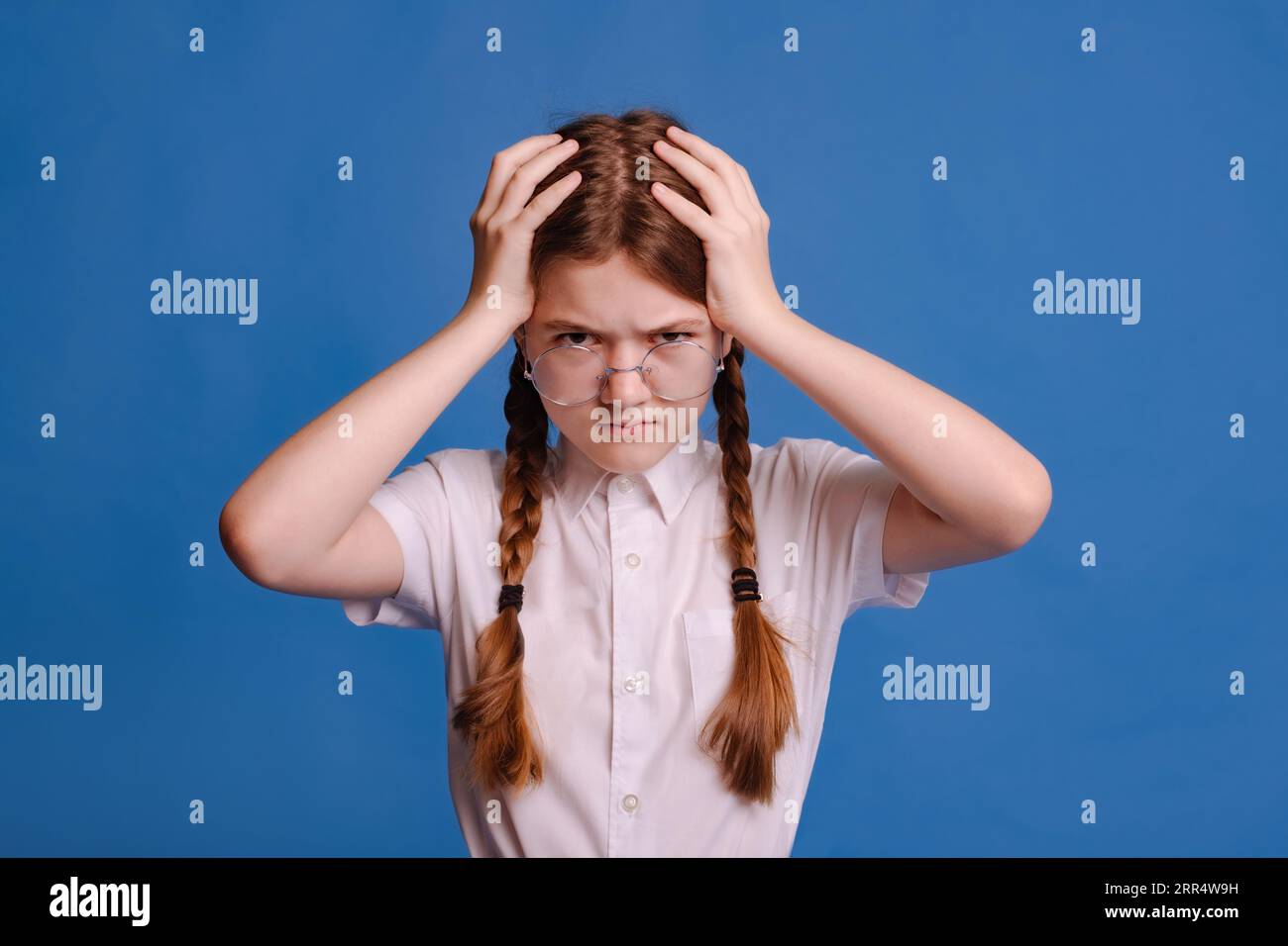 schoolgirl in round glasses on blue background confuse and wondering about question. uncertain with doubt, thinking with hand on head. pensive concept Stock Photo