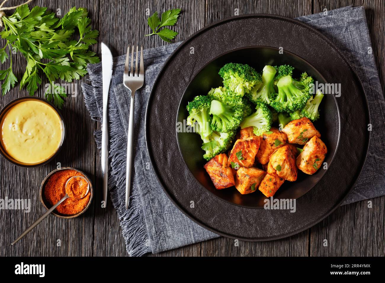 fried salmon bites with cooked broccoli florets in black bowl on dark wooden table with sriracha mayo sauce, horizontal view from above Stock Photo