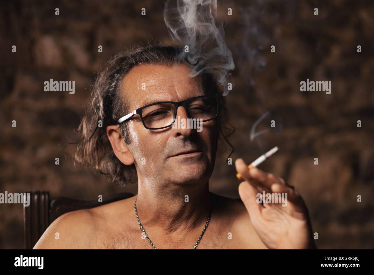 An older man, about 60 years old, of Serbian nationality with long hair and glasses, thoughtfully smokes a cigarette on a dark background. Artist, cre Stock Photo