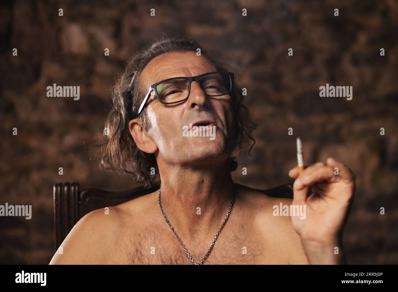 An older man, about 60 years old, of Serbian nationality with long hair and  glasses, thoughtfully smokes a cigarette on a dark background. Artist, cre  Stock Photo