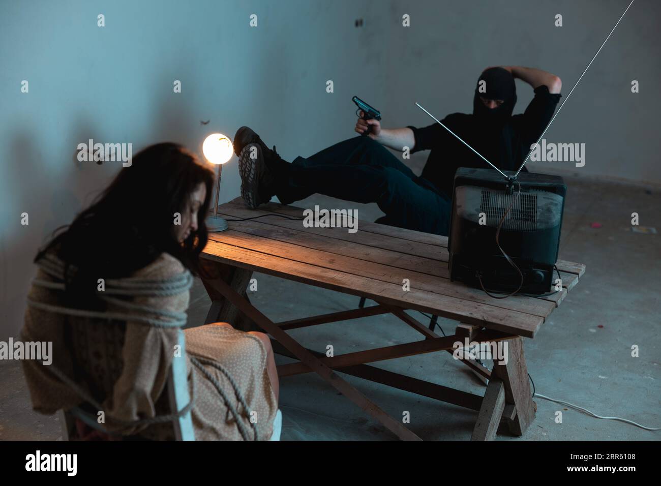 Kidnapper in balaclava sitting in front of the kidnapped woman and holding a gun while watching TV. Crime, kidnapping, violence concept Stock Photo