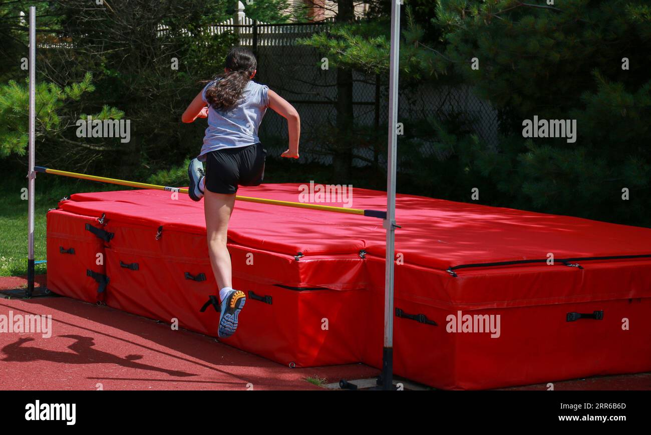Rear view of a teeange girl jumping over a yellow high jump bar on to red high jump mats. Stock Photo
