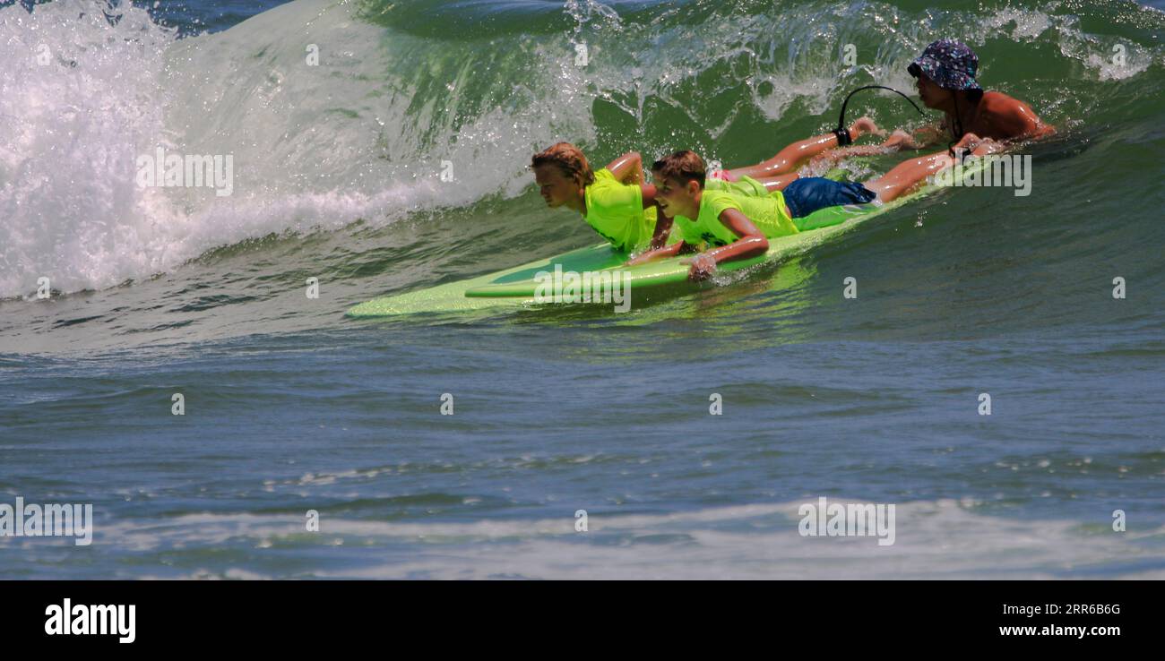 Gilgo Beach, New York, USA - 25 July 2023: Surf camp counselor holding two boys in neon green shirts on their surfboard helping then catch a wave to g Stock Photo