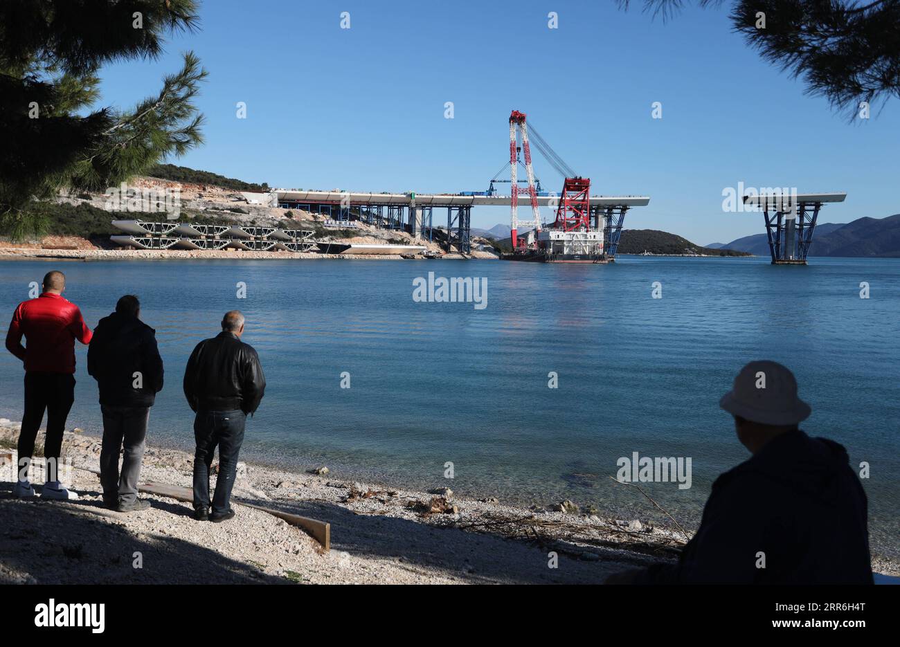 210215 -- KOMARNA CROATIA, Feb. 15, 2021 -- People watch as a crane carries a steel box girder to connect the Croatian mainland with the first sea-based pillar of the Peljesac Bridge near Komarna, Croatia, Feb. 15, 2021. The construction of the Peljesac Bridge, the biggest infrastructure project in Croatia, will be completed in June 2022, Prime Minister Andrej Plenkovic announced on Monday after visiting the site. The 2.4-kilometer-long Peljesac Bridge connects Croatia s southernmost Dubrovnik-Neretva County to the rest of the mainland, giving the southeastern European country a continuous lan Stock Photo