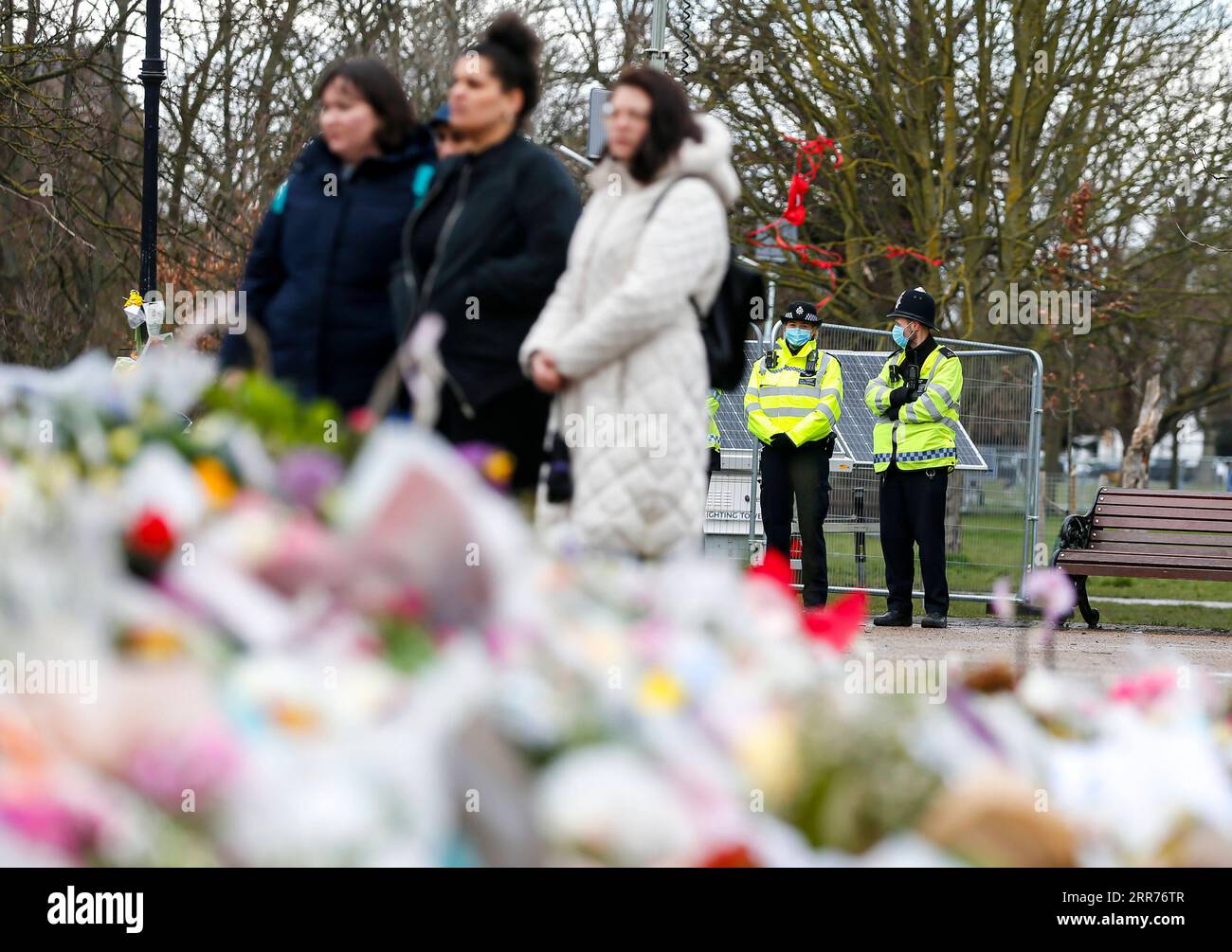 210316 -- LONDON, March 16, 2021 -- People stand next to floral tributes at the bandstand on Clapham Common to mourn for Sarah Everard while police officers stand guard in London, Britain, on March 15, 2021. A serving Metropolitan police officer on March 13 appeared in court in London after being charged with the kidnap and murder of a 33-year-old woman. Wayne Couzens, 48, was arrested after Sarah Everard, a marketing executive, went missing while walking home from a friend s apartment in south London on March 3. Everard s death has caused widespread concern in Britain about women s safety, wi Stock Photo