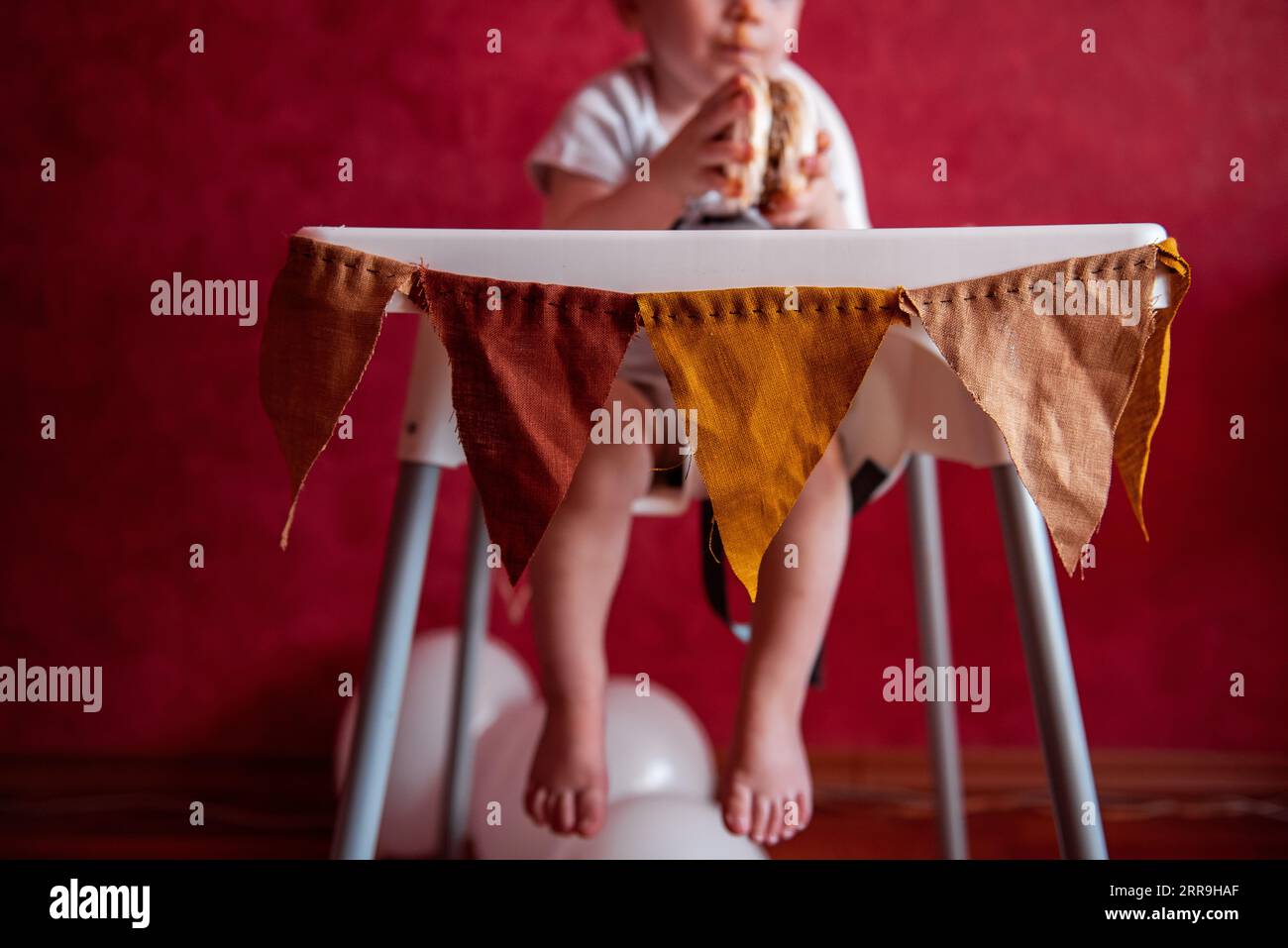 Faceless portrait of baby sitting in feeding chair, festively decorated with fabric pennants for the birthday party. On red textural background with b Stock Photo