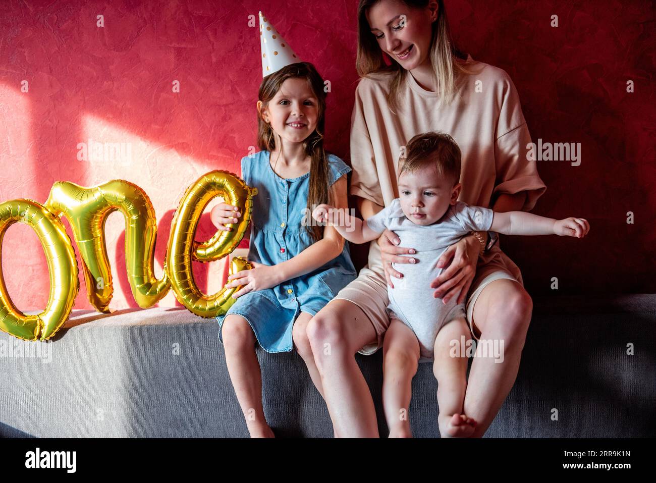 Young mother sits on sofa with small children near red wall on gray sofa. Family with one parent celebrates the first year of baby. Older sister is ho Stock Photo